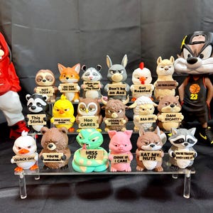 Pode incluir: Uma coleção de figuras de animais engraçados, cada uma segurando uma placa com uma frase engraçada. As figuras incluem uma preguiça, uma raposa, um guaxinim, um coelho, uma galinha, uma lhama, um pato, um urso, uma cobra, um porco, um esquilo e um guaxinim.