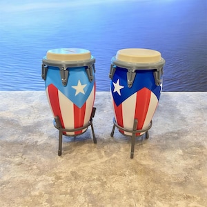 May include: Two conga drums with the Puerto Rican flag design. The drums have a blue top and a beige drumhead. The body of the drums is painted with the Puerto Rican flag, featuring red and white stripes, a blue triangle, and a white star. The drums are supported by a gray metal stand.