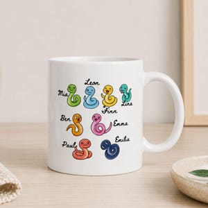 Könnte beinhalten: Ein weißer Keramikbecher mit einer bunten Illustration von acht Cartoon-Schlangen in verschiedenen Farben, darunter Grün, Blau, Gelb, Orange, Pink und Dunkelblau. Jede Schlange ist mit einem Namen beschriftet: Mia, Leon, Finn, Lina, Ben, Emma und Paul.