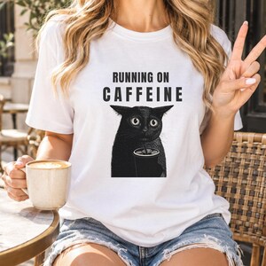 Op de afbeelding: Een wit t-shirt met korte mouwen en een zwarte afbeelding van een kat met grote ogen die een kop koffie vasthoudt. Boven de kat staat de tekst RUNNING ON CAFFEINE in dikke zwarte hoofdletters. Het shirt heeft een ronde hals en pasvorm.