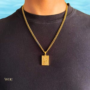 May include: Gold-colored rectangular pendant necklace with a detailed design, suspended from a matching curb chain. The necklace is worn against a black shirt. The word "WOU" is visible in the lower left corner.