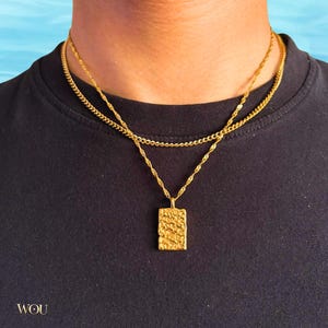 May include: A layered gold-tone necklace set featuring a shorter, thin curb chain and a longer, twisted rope chain. The rope chain holds a rectangular pendant with a textured, hammered surface. The word WOU is printed in the bottom left corner.