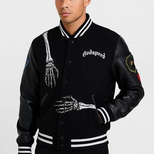 Könnte beinhalten: Schwarze Collegejacke mit weißem Besatz und schwarzen Lederärmeln. Verfügt über eine Skeletthand-Grafik und das Wort "Godspeed" in weißer Schrift. Enthält einen Aufnäher auf dem Ärmel.