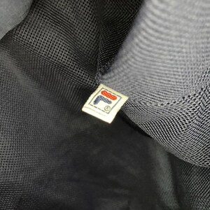 May include: Close-up of a black mesh fabric with a small rectangular tag. The tag features the FILA logo in red, white, and blue, with the brand name and a registered trademark symbol.