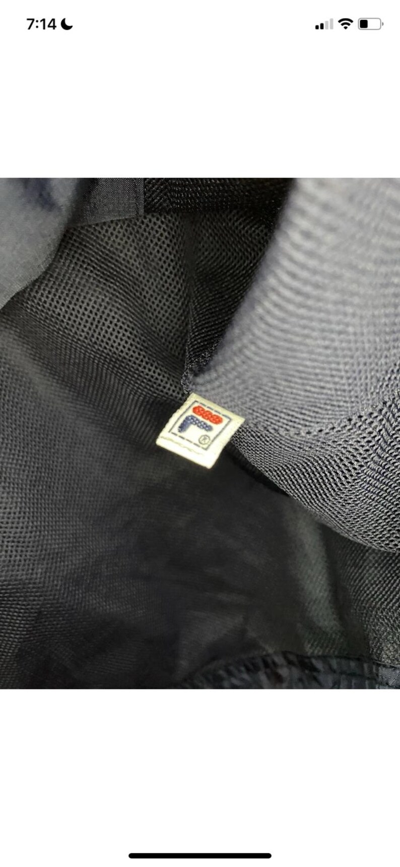 May include: Close-up of a black mesh fabric with a small rectangular tag. The tag features the FILA logo in red, white, and blue, with the brand name and a registered trademark symbol.