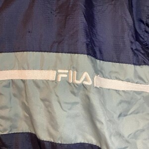 May include: Close-up of a navy blue and light blue Fila jacket. The jacket features a light blue stripe across the chest with the Fila logo in white. The jacket is made of a shiny, possibly nylon, material.