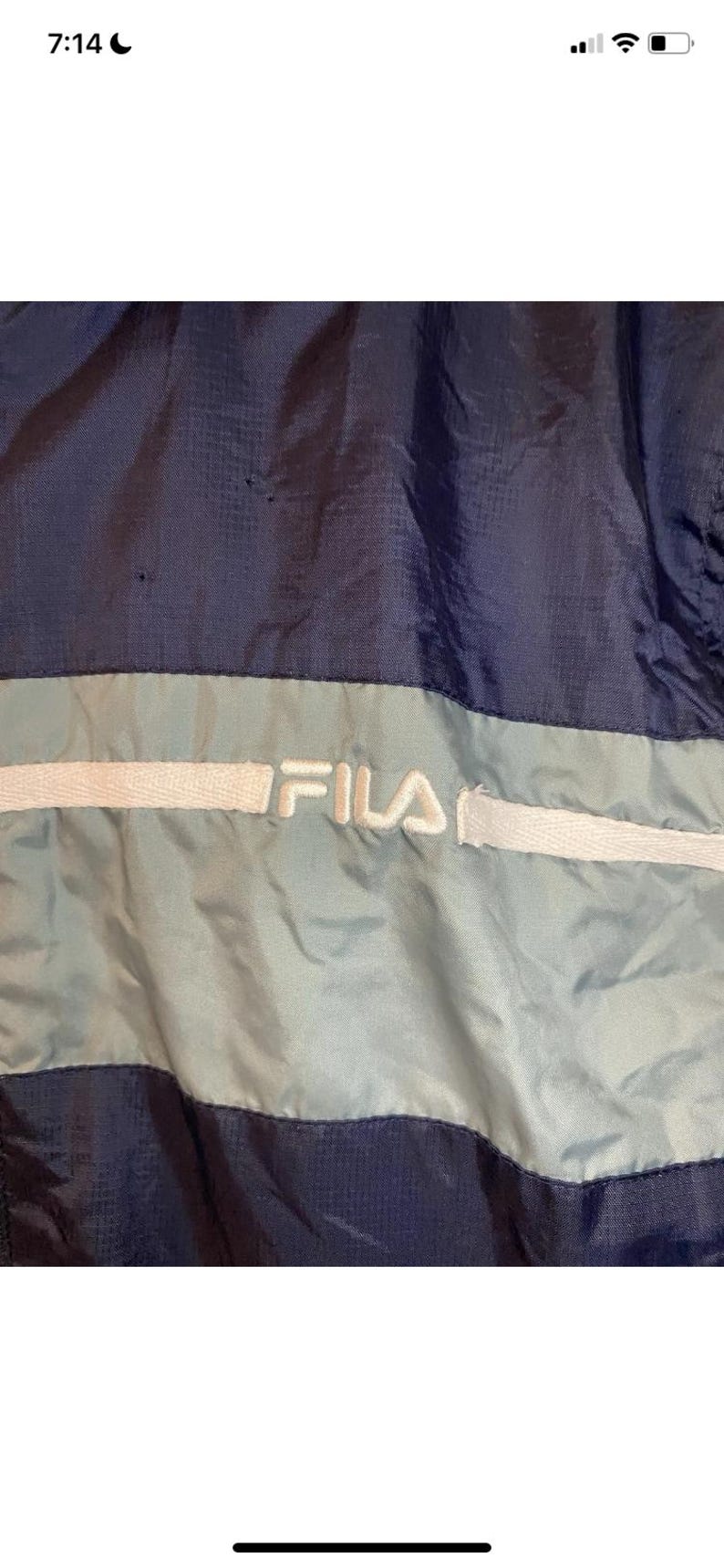 May include: Close-up of a navy blue and light blue Fila jacket. The jacket features a light blue stripe across the chest with the Fila logo in white. The jacket is made of a shiny, possibly nylon, material.