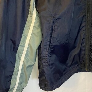 May include: Navy blue windbreaker jacket with a light blue and white stripe detail on the sleeve. The jacket has a zipper closure and a pocket. The material appears to be a lightweight nylon or similar fabric.