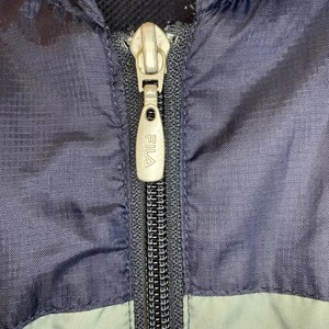 May include: Close-up of a navy blue and light blue jacket with a silver zipper. The zipper pull has the brand name "FILA" printed on it. The jacket appears to be made of a lightweight material.