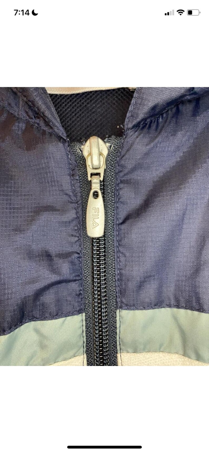 May include: Close-up of a navy blue and light blue jacket with a silver zipper. The zipper pull has the brand name "FILA" printed on it. The jacket appears to be made of a lightweight material.