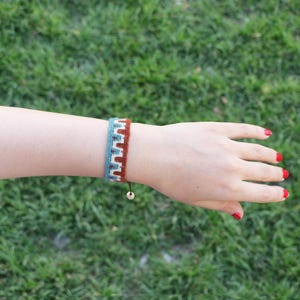 May include: A woven bracelet with a geometric pattern in shades of blue, white, and brown. The bracelet is on a person's wrist, with a wooden bead and adjustable string closure. The background is green grass.