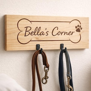 Pode incluir: Placa de madeira com o texto "Bella's Corner" e uma pegada, emoldurada por um osso. Dois ganchos de metal preto seguram uma trela castanha e uma azul. A placa está montada numa parede branca.