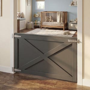 Op de afbeelding: Een grijze houten babyhek met een klassiek X-ontwerp, geïnstalleerd in een deuropening. De hek heeft zilveren scharnieren en een grendel. De achtergrond toont een kinderkamer met een wieg, vloerkleed en planken.