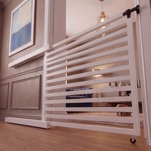 May include: A white, slatted baby gate with a rolling wheel at the bottom. The gate is installed in a doorway, with a black latch on the door frame. The gate is made of wood and is designed to keep children safe.