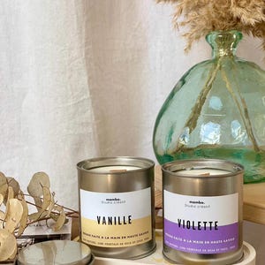 Peut inclure: Deux bougies en métal avec des étiquettes indiquant "Vanille" et "Violette". Les bougies sont posées sur un plateau décoratif. L'arrière-plan comprend un vase en verre avec des plantes séchées et un rideau blanc.