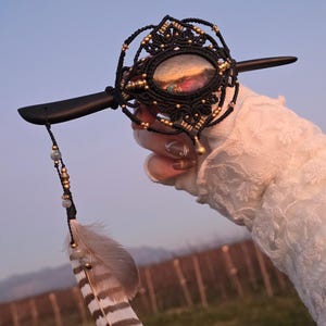 Könnte beinhalten: Handgefertigtes Haaraccessoire mit schwarzem Makramee-Design, goldenen Akzenten und einem zentralen Stein. Ein dekorativer Feder- und Perlenanhänger hängt an dem Holzstab. Das Accessoire vor einem verschwommenen Hintergrund.