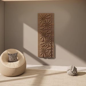 Könnte beinhalten: Ein Wandkunstwerk aus Holz mit einem Blumendesign aus fünf geschnitzten Blumen. Die rechteckige Kunst ist an einer neutralfarbenen Wand angebracht. Ein beiger Sitzsack mit einem gemusterten Kissen und ein graues Knotenkissen befinden sich im Vordergrund.