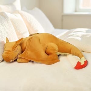 May include: A plush toy shaped like a dragon with orange-yellow skin, folded wings, and a long tail ending in a red and cream-colored flame tip. The creature is depicted in a sleeping position with closed eyes and a curved mouth line on its face.