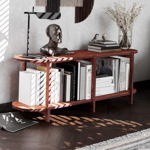 May include: A dark wood, two-tiered console table with rounded edges. The top shelf holds a sculpture, books, and a lamp. The lower shelf is filled with books. A vase of dried flowers sits on the top shelf. The table is in a living room setting.
