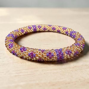 Peut inclure: Un bracelet de perles avec un motif doré et violet. Le bracelet est composé de petites perles rondes, avec des perles dorées formant la base et des perles violettes créant un motif floral. Le bracelet est circulaire et repose sur une surface en bois clair.