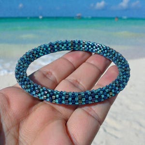 May include: A beaded bracelet in shades of blue and teal. The bracelet is made of small, evenly spaced beads, creating a textured, cylindrical shape. The bracelet is held against a backdrop of a beach with turquoise water and a clear blue sky.
