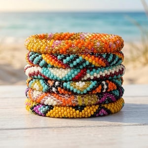 Pode incluir: Uma pilha de seis pulseiras de contas em várias cores. As pulseiras são feitas de pequenas contas coloridas e apresentam padrões de laranja, amarelo, turquesa, preto, branco e roxo. As pulseiras estão empilhadas sobre uma superfície branca com uma praia e o oceano ao fundo.