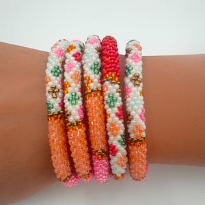 May include: A collection of beaded bracelets in various colors. The bracelets feature a mix of orange, pink, white, and red beads, with floral and geometric patterns. The bracelets are approximately 2.5 inches in diameter.