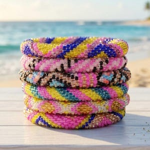 May include: A stack of six beaded bracelets in various colors. The bracelets are stacked on top of each other, with a blurred ocean background. The bracelets feature pink, yellow, blue, and black beads, creating a colorful and textured appearance.