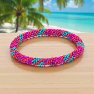 Può includere: Un braccialetto di perline colorate nei toni del rosa, viola e turchese. Il braccialetto è composto da piccole perline rotonde disposte a spirale. Lo sfondo presenta una scena di spiaggia tropicale.