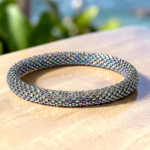 Op de afbeelding: Een kralenarmband met zilveren en iriserende kleuren. De armband is gemaakt van kleine, dicht geweven kralen, waardoor een textuur ontstaat. De armband is cirkelvormig en ligt op een lichtgekleurd houten oppervlak.
