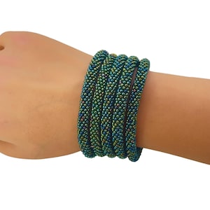 Puede incluir: Seis pulseras de cuentas en tonos turquesa, verde y azul. Las pulseras están tejidas apretadamente, creando una superficie texturizada. Las cuentas tienen un brillo iridiscente sutil, agregando profundidad a la joyería. Las pulseras se usan en la muñeca.
