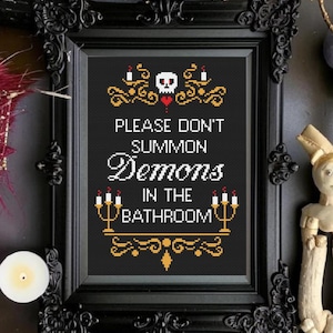 Peut inclure: Une œuvre d'art au point de croix encadrée avec le texte "PLEASE DON'T SUMMON DEMONS IN THE BATHROOM". Le motif comprend un crâne, des bougies et des éléments décoratifs dorés et rouges. Le cadre est noir et orné.