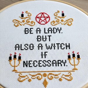 Pode incluir: Bordado em ponto cruz num bastidor de madeira. O tecido branco apresenta o texto "BE A LADY. BUT ALSO A WITCH IF NECESSARY." em preto, com detalhes dourados e vermelhos. Elementos decorativos incluem um pentagrama e castiçais.