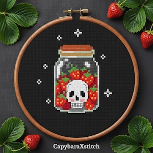 Pode incluir: Design de ponto cruz com um frasco cheio de morangos vermelhos e uma caveira. O design é definido contra um fundo preto dentro de um bastidor de bordado de madeira. Morangos frescos e folhas verdes circundam o bastidor.