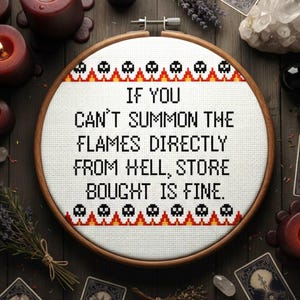 May include: Cross-stitch embroidery in a wooden hoop with the text "IF YOU CAN'T SUMMON THE FLAMES DIRECTLY FROM HELL, STORE BOUGHT IS FINE." The design includes skulls and flames. The image also features candles, tarot cards, and dried lavender.