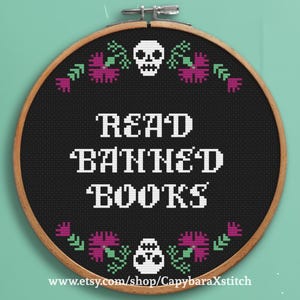 Pode incluir: Um bastidor de bordado circular de madeira contendo tecido preto com o texto READ BANNED BOOKS bordado em ponto cruz branco. O design apresenta duas caveiras brancas e motivos florais rosa e verdes acima e abaixo do texto.