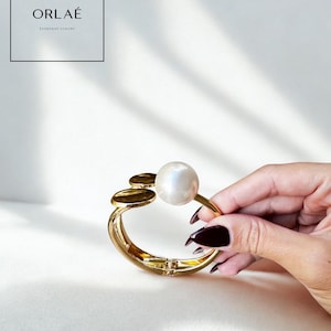 May include: A gold bracelet featuring a large, round, white pearl and three smaller gold discs. The bracelet is displayed against a white backdrop. The brand name "ORLAÉ" and the tagline "EVERYDAY LUXURY" are visible in the top left.