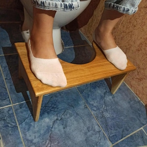 May include: A wooden toilet stool with a light brown finish. The stool is rectangular with angled legs and is designed to be placed in front of a toilet. The stool is approximately 12 inches long.