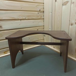May include: A brown wooden stool with a unique curved design. The stool has four angled legs and a horizontal support bar. The top surface features a curved cutout, and the wood grain is visible. This piece is suitable for home decor.