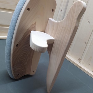 Könnte beinhalten: Ein Bügelbrett aus Holz mit einer grau gepolsterten Oberfläche. Das Brett hat ein gebogenes Design mit einer Holzstützstruktur und einer kleinen, angebrachten Ablage. Das Holz hat eine natürliche Oberfläche.