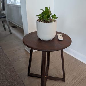 May include: A small, round, dark brown wooden side table with a plant in a white, ribbed ceramic pot. A white remote control rests on the table's surface. The table has a three-legged base.