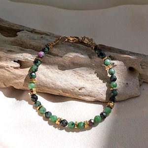 Puede incluir: Una pulsera de cuentas con pequeñas piedras facetadas verdes y gris oscuro intercaladas con diminutos espaciadores metálicos de tono dorado. La pulsera incluye un cierre de mosquetón dorado y una pequeña piedra redonda de color rosa oscuro.