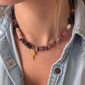 Puede incluir: Un collar de cuentas con un colgante dorado. El collar presenta una mezcla de cuentas rosas, negras y blancas. El colgante dorado tiene un diseño de rostro. El collar se lleva sobre una camisa vaquera.