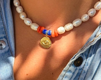 Könnte beinhalten: Eine Perlenkette mit einem goldenen Anhänger mit Augenmotiv. Die Kette hat einen Abschnitt aus orangefarbenen und blauen Perlen. Die Kette wird über einem Jeanshemd getragen.