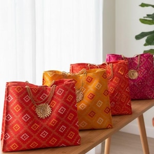 May include: Five colorful tote bags with a geometric pattern in shades of red, orange, and yellow are arranged on a wooden surface. Each bag has rope handles and a decorative gold-colored embellishment.