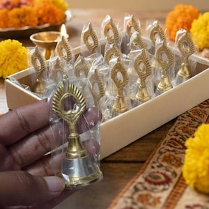 Puede incluir: Una caja de pequeñas campanas decorativas doradas, cada una envuelta individualmente en plástico transparente. Las campanas tienen un diseño único con una abertura circular en la parte superior. La imagen también muestra flores amarillas y naranjas.