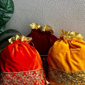 May include: Three velvet drawstring bags in shades of red and gold. The bags have gold trim and drawstrings. The bags are likely for gifts or special occasions. The bags are on a decorative surface.