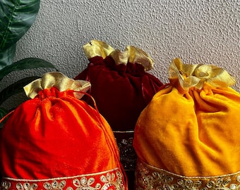 May include: Three velvet drawstring bags in shades of red and gold. The bags have gold trim and drawstrings. The bags are likely for gifts or special occasions. The bags are on a decorative surface.