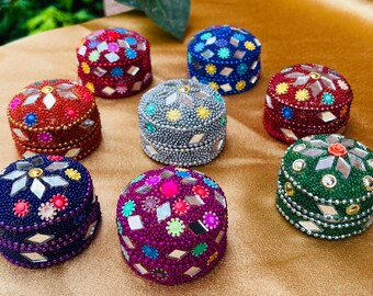 May include: A collection of small, round, decorative boxes. Each box is covered in colorful beads and mirrored accents. The boxes are in various colors, including red, blue, green, purple, and silver. The boxes are arranged on a gold-colored surface.