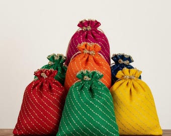 Könnte beinhalten: Eine Sammlung farbenfroher Beutel mit Kordelzug in verschiedenen Farbtönen, darunter Rot, Grün, Orange, Gelb, Pink und Blau. Jeder Beutel hat ein diagonales Streifenmuster und einen dekorativen, perlenbesetzten Verschluss. Die Beutel sind auf einer Holzoberfläche angeordnet.